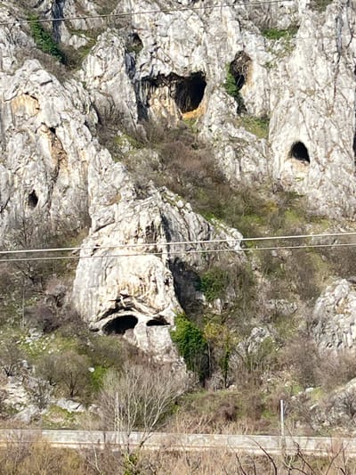 This image depicts a rugged, light-grey limestone rock face located in Lyutibrod, Bulgaria, under bright, direct sunlight. The rock formation is heavily fractured and eroded, presenting numerous cavities and overhangs.
In the upper half of the image, multiple dark, irregular cave openings are visible. A particularly prominent, large, dark opening, roughly heart-shaped or skull-like in appearance, is located towards the upper-middle right. Several smaller, rounded, or oval openings are scattered across the upper and mid-sections of the rock face.
Dominating the lower-middle section is a distinct natural rock formation that strikingly resembles a large human skull or face. This formation features two deeply recessed, dark, and somewhat triangular hollows appearing as 'eye sockets,' with a broad, shadowed overhang beneath them forming a 'brow.' Below these, a wide, elongated dark cavity or overhang suggests a 'mouth.'
Sparse vegetation, primarily deciduous trees and shrubs with bare branches, is present on the lower slopes and within crevices of the rock face. Patches of evergreen growth are also visible in sheltered areas. Two thin, parallel horizontal wires, likely utility lines, traverse the mid-section of the image, partially obscuring the rock. At the very bottom of the frame, a horizontal segment of a paved surface or railway track is visible, indicating proximity to human infrastructure at the base of the rock formation.