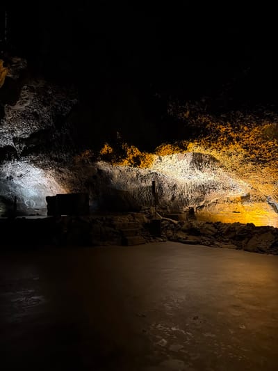 This image depicts a section of the Cueva de los Verdes, a lava tube located in Haría, Spain. The left side of the image features a rugged, textured rock wall illuminated with amber and white light. The right side shows a smooth, flat wall, possibly man-made, in shadow. A series of wooden beams and supports are anchored into the rock wall near the center of the frame. The environment is dark, suggesting the underground location of the cave.