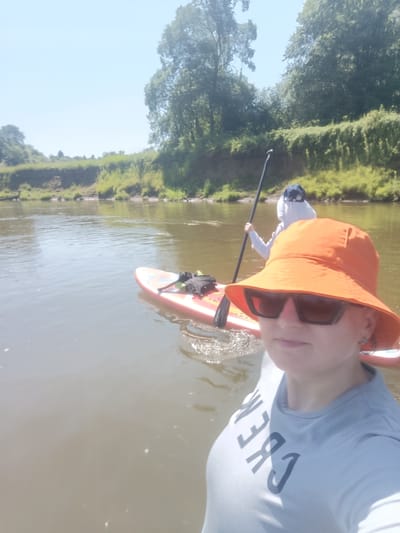 In the city of Berkuty, Russia, two individuals are paddleboarding on a wide, calm river under a bright, clear sky. The person in the foreground, a woman wearing an orange bucket hat and sunglasses, is taking a selfie and is positioned on a paddleboard. She is wearing a light blue t-shirt with dark lettering that reads "CREW". Behind her, another person is also on a paddleboard, wearing a light-colored hooded jacket and a dark baseball cap. This individual is holding a paddle and appears to be moving through the water. The paddleboards are red and white. The riverbanks are lined with lush green trees and vegetation, with a gentle slope leading down to the water. The water itself is murky brown, reflecting the sunlight.