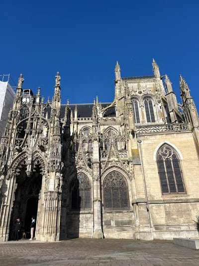 The image displays the ornate Gothic facade of a large stone church in Louviers, France, captured from a low angle under a clear, cloudless blue sky. The building's exterior features extensive intricate stone tracery, multiple pointed arches, numerous sculpted figures within niches, pinnacles, and spires. White scaffolding encloses a section of the facade on the far left, indicative of ongoing construction or renovation. The main entrance, located on the left, consists of deeply recessed, highly decorated pointed arch portals. Three individuals are observed near the leftmost portal: one subject in a dark jacket stands beside a light-colored dog on a leash, another subject in lighter clothing is positioned further within the doorway, and a third subject is partially visible. The immediate foreground comprises a paved surface of light-toned rectangular and square stones. Strong sunlight illuminates the scene, casting distinct shadows across the architectural details.
