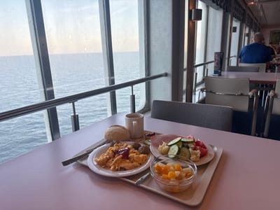 The image shows a meal served on a tray on a pink table in front of a large window overlooking the ocean. The meal consists of scrambled eggs, meatballs, a bread roll, sliced ham, a salad with cucumbers, tomatoes, and cauliflower, a small bowl of fruit salad, and a mug of coffee with a small dish of jam. A fork and knife are placed on the tray. In the background, through the window, the calm ocean and sky are visible. Inside the dining area, tables and chairs are arranged, and a man in a blue shirt is seated at a table in the distance. The overall environment appears to be a dining room or cafeteria with a view of the sea.