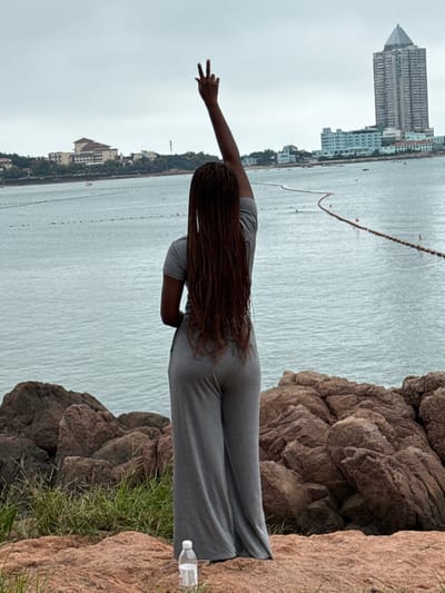 A female individual is observed from behind, standing on a rocky incline overlooking a body of water under an overcast sky. Her hair consists of long, reddish-brown box braids extending down her back. She wears a short-sleeved gray top and loose-fitting gray wide-leg pants. Her right arm is raised vertically, with the index and middle fingers extended upwards. At her feet, a clear plastic water bottle with a white cap rests on the reddish-brown rock surface.
The midground features a calm sea with a long, undulating floating barrier marked with orange buoys. In the distant background, the urbanized coastline of Shinan District, Qingdao, China, is visible. To the left, several buildings with light facades and red-tiled roofs are nestled among greenery. To the right, a tall, modern skyscraper with a dark, pointed roof (identified as Haiwang Mansion) stands prominently, flanked by several lower-rise structures along the water's edge. The foreground consists of rough, reddish-brown rock formations interspersed with patches of dry green grass.