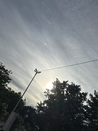 The image shows a low-angle view of a partially cloudy sky, a power line, and a street light pole. The sun is partially obscured by trees on the right side of the image. The street light pole is diagonally oriented, starting from the lower right and extending towards the upper center. The leaves of the trees are silhouetted against the bright sky. There is a box-like structure near the base of the streetlight. There are no visible people in the image. The exact location within Glojene, Bulgaria cannot be determined from the given image.