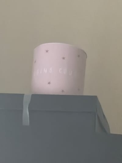 The scene is an interior view within the city of Lurgan, United Kingdom. A light pink cylindrical object, approximately a candle holder or decorative pot, is positioned centrally on top of a dark grey, rectangular object, possibly a box or flat-topped container. The pink object features multiple small, darker pink star patterns and partially visible white text reading "SINS COU" on its surface. A light-colored ribbon or strap hangs vertically over the front edge of the dark grey object. The background is a plain, light beige or off-white wall or surface. No individuals or discernible actions are present.