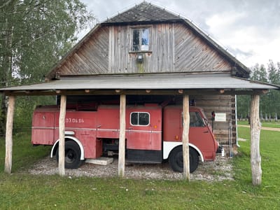 A red vintage fire truck is parked inside a weathered wooden structure that resembles a barn or shed. The fire truck has the number "03 04 64" painted on its side. The structure has a sloped roof covered in grey shingles, and wooden beams support a lower, overhanging roof. The walls are constructed from vertical wooden planks. A single window is visible on the upper part of the main structure. The ground around the fire truck is covered with gravel, and lush green grass surrounds the structure. In the background, trees and a cloudy sky are visible. A sign on the wooden wall of the structure reads "STAR A-25/A-26 rok produkcji 1971." This scene is located in Sielachowskie, Poland.