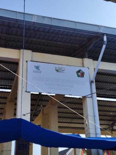 A sign reads "Pasar Rakyat Beureundeun, Kabupaten Pidie, DAKA TP. 2022" in light gray lettering against a white background. The sign also features logos of "Kementerian Perdagangan" and "Pasar Rakyat," alongside a colorful seal. The sign is mounted on a building support column under a metal roof structure. Below, a blue tarp partially obscures the view.