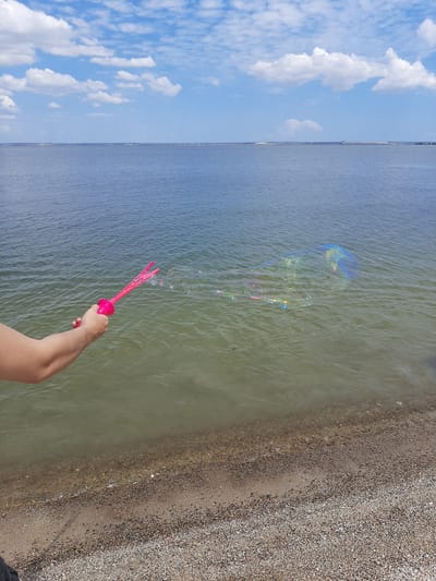 In Sukholuzhzhia, Ukraine, on a bright, partly cloudy day, a person's arm and hand are visible on the left side of the frame. The hand is holding a pink bubble wand with three loops, from which a trail of iridescent bubbles is floating on the water's surface. The bubbles are large and reflect the light, creating a rainbow effect. The scene takes place by the water's edge, with a pebbly beach in the foreground and the calm, blue-green water extending to the horizon. The sky is a vibrant blue with scattered white clouds. The overall impression is one of a leisurely outdoor activity by a body of water.