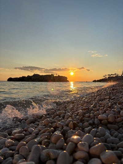 The image captures a serene sunset scene on a pebbled beach in Dobra Voda, Montenegro. The foreground is dominated by smooth, water-worn pebbles glistening under the warm glow of the setting sun. Small waves gently lap the shore, creating splashes of water that catch the light. In the mid-ground, the calm sea reflects the vibrant orange and yellow hues of the sunset. Across the water, a rocky island with a few silhouetted structures stands against the colorful sky. The horizon is marked by a sliver of land with several umbrellas and indistinct figures, suggesting a populated area. The sky transitions from a pale blue at the top to a warm gradient of orange and yellow near the horizon, with a few wispy clouds. The overall impression is one of tranquility and natural beauty.