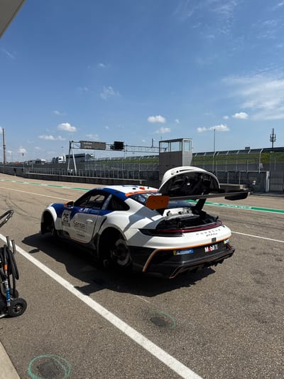 The image displays a stationary Porsche 911 GT3 Cup race car, numbered 25, within the pit lane area of the Sachsenring circuit in Hohenstein-Ernstthal, Germany. The scene is illuminated by bright daylight under a clear blue sky with sparse white clouds.
The race car exhibits a white, blue, and orange livery with prominent "Sensei I", "Mobil 1", and "Michelin" sponsor markings. Both the left front and left rear wheels are absent, exposing the brake discs and wheel hubs. The rear engine cover is open, revealing internal mechanical components of the engine bay. A large orange rear wing is affixed to the car.
The ground surface consists of asphalt, marked with white lines defining the pit lane. Two green chalk circles are visible on the asphalt in the immediate foreground. To the far left, a black utility cart equipped with hoses and connectors is partially visible.
In the background, an overhead gantry spans the track, supporting a digital display board that indicates "20.08.2023" and "10:48:45". Below the display, a set of track traffic lights is visible. Adjacent to the track, a multi-level pit building or control tower stands. Safety barriers and a line of parked support vehicles are present further in the background. No individuals are depicted in the scene.