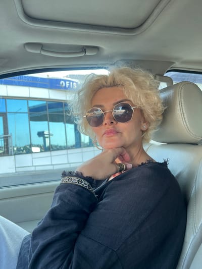 A woman with short blonde curly hair and sunglasses is seated in the passenger seat of a car. She is wearing a dark top with embroidered detailing on the sleeves and a ring on her right hand. Her left hand is resting on her chin, and her head is tilted slightly. The interior of the car, including the headrest and ceiling handle, is visible. Outside the car window, a blue-tinted building with "OFIS" signage is reflected in the glass. This scene is located in the city of Khirdalan, Azerbaijan.