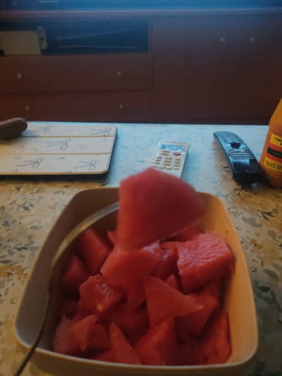 The image captures an indoor scene, likely a domestic setting, with a focus on a bowl of chopped watermelon placed on a patterned tablecloth. In the background, a dark wooden piece of furniture with drawers is visible, along with a glimpse of what appears to be an electronic device. Several remote controls are present on the table, alongside a bottle of what seems to be hot sauce. There are also three coasters with a design resembling scissors and the number 8. The lighting suggests an indoor environment, possibly a home in Figueres, Spain, though no specific landmarks or indicators of this location are present in the image itself. The scene is static, with no people or animals depicted.