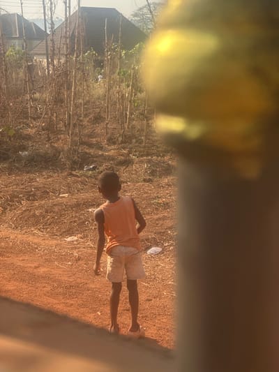 A young individual stands facing away from the viewer on reddish-brown dirt, partially obscured by a blurred, golden-yellow object on the right side of the frame. The individual wears an orange sleeveless top, light-colored shorts, and light-colored open-toed footwear. The ground around the individual is a mix of bare soil and scattered dry vegetation, with visible pieces of white debris. In the midground, numerous thin, dry plant stalks, interspersed with some green foliage, cover a substantial area. Further in the background, two buildings with dark, sloped roofs are discernible, with one appearing light-colored. Distant trees line the horizon beneath a bright, hazy sky. The scene is illuminated by direct sunlight from the left, casting subtle shadows. This location is identified as Umuoye, Nigeria.