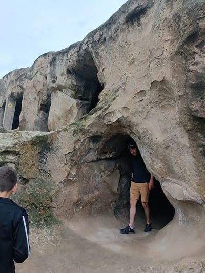 The image depicts two individuals exploring the cave city of Uplistsikhe, Georgia. In the foreground, a person with short brown hair, wearing a black jacket with white stripes on the sleeves, is viewed from the back, facing the caves. To the right, a man wearing a black t-shirt, a black baseball cap, and tan shorts stands partially inside a cave opening, looking towards the camera. The environment is characterized by large rock formations with natural cave entrances, suggesting an ancient historical site. The ground is sandy and uneven. The sky is overcast and light in color.
