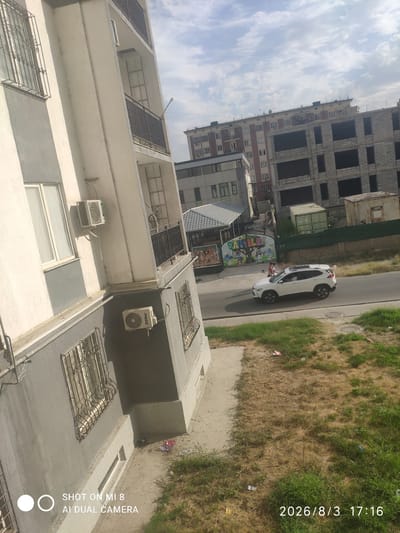 This is a high-angle, full-frame, eye-level shot of a street scene in Charjuy, Uzbekistan, on August 3, 2026, at 17:16. In the foreground, a portion of a multi-story building is visible on the left, with balconies and windows. One window has ornate metal bars. Two air conditioning units are mounted on the building's exterior. The building appears to be under construction or renovation in some areas.
A white SUV is driving on the paved road in the middle ground. Behind the SUV, there is a low-rise building with a patterned roof, and beyond that, a larger, multi-story building that appears to be under construction, with exposed concrete framing and dark window openings. Further in the background, another building with many windows is visible. There is some scattered debris on the ground, including grass, dry vegetation, and some litter. The sky is partly cloudy. The overall scene depicts an urban environment with a mix of older and newer construction.