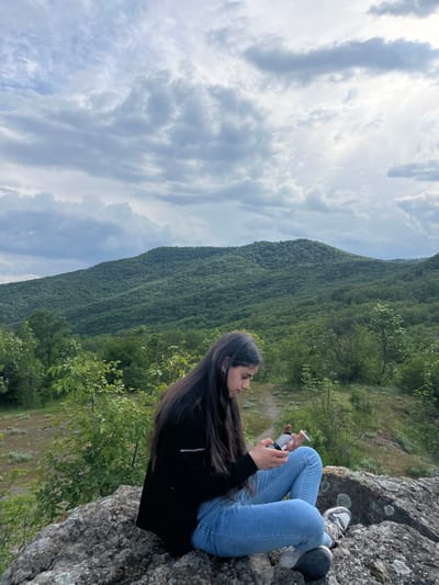 A woman is sitting on a rocky outcrop in a mountainous, wooded landscape. She is wearing a black jacket, light blue jeans, and grey sneakers. Her long dark hair falls over her shoulder. She is looking at a small object in her hands. A mountain covered in dense forest is in the background. The sky is filled with white and grey clouds. The location is likely near Kamchia, Bulgaria, based on the landscape features.