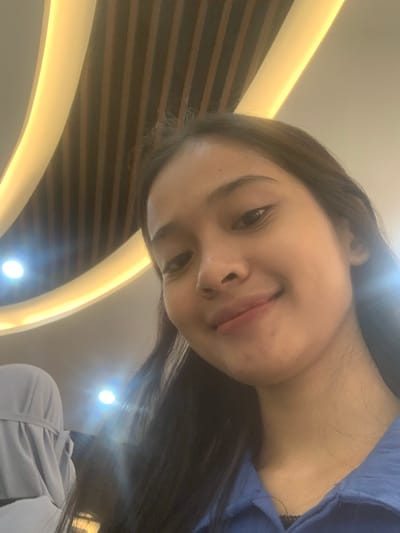 The image is a close-up, diagonally oriented portrait of a young person, likely female, captured indoors in Samarinda, Indonesia. The individual, who has long dark hair and fair skin, is looking directly at the camera with a soft, slightly playful expression, featuring a gentle smile and subtly parted lips. She is wearing a blue top, visible over her shoulder and chest.

The background suggests a modern interior. Dominant features include curved, illuminated architectural elements that appear as bright, glowing arcs on a light-colored surface, possibly a ceiling or wall. In contrast, dark horizontal wooden slat panels are also visible, adding texture to the background. Several bright, circular light sources or reflections are scattered throughout the scene, creating lens flares and contributing to the well-lit environment. A glimpse of another person or object, possibly wearing a light grey or white garment, can be seen in the lower right portion of the frame. The overall impression is a candid, selfie-style photograph taken in a contemporary setting. No visible text is present in the image.