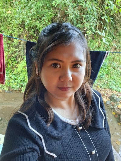 A brunette woman with light skin gazes at the camera with brown eyes and a faint smile. She is wearing a dark, knitted sweater with white trim and pearl-like buttons. Lush green foliage and some bare branches surround her. The background also reveals what appears to be a concrete or stone surface, suggesting an outdoor setting. Part of a dark object with a reddish trim is visible near the bottom left corner.