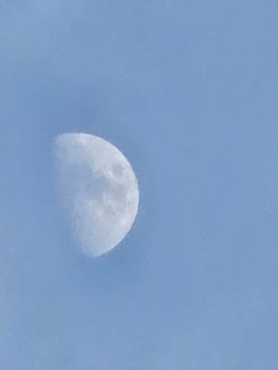 The image shows a crescent moon against a pale blue sky. The moon is not fully illuminated, with about half of it visible. The surface of the moon is textured, suggesting craters and other lunar features, but the details are somewhat blurred. The sky is a uniform light blue, with no clouds or other celestial bodies visible. The overall impression is of a clear day or early evening in Gumi-si, South Korea, where the moon is visible in daylight or twilight. There is no text or identifiable human activity in the image.