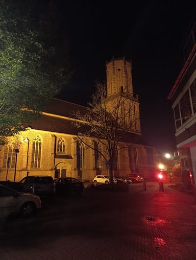 At night in Emmerich on the Rhine, Germany, a brick church with a tall tower stands illuminated. A bare tree is positioned in front of the church, which is flanked by parked cars. A red traffic signal is visible on the right side of the image, along with other buildings and streetlights. The ground is bricked, and reflective in spots. Some leafy trees are on the left.
