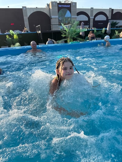 The image shows a group of people in a swimming pool. In the foreground, a young girl with braided hair is splashing in the water, smiling at the camera. Behind her, several other people are visible in the pool, including an adult man with a shaved head wearing a necklace. The background features a building with large arched windows, palm trees, and umbrellas, suggesting an outdoor recreational area. A sign with the text "termoland" is visible on the building. The sky is clear and bright, indicating it is daytime. The overall scene appears to be a sunny day at a water park or resort.