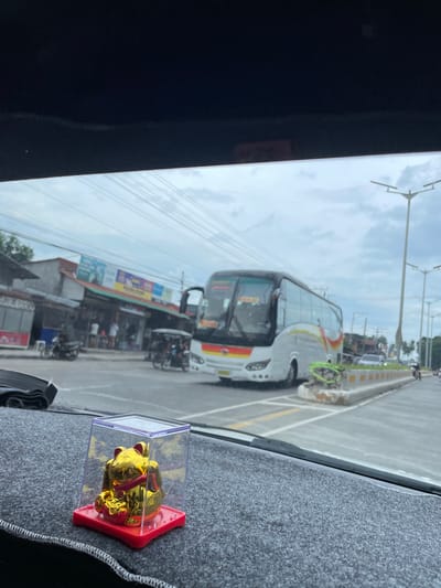 The image, taken from inside a vehicle, shows a view through the windshield of a bus on a road in Santa Cruz, Philippines. A small golden lucky cat figurine in a plastic display case sits on the dashboard. Outside, the bus is in the right lane, with streetlights and power lines visible overhead. Buildings and motorcycles line the street.