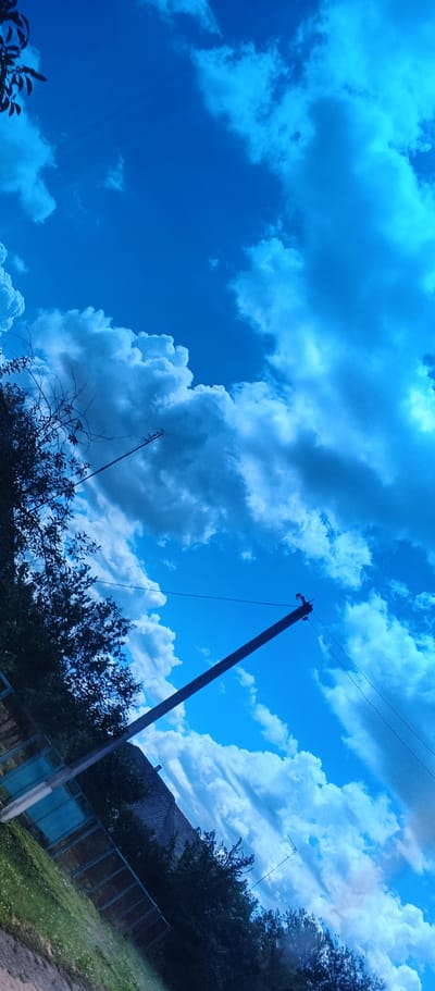 The image depicts a bright blue sky filled with fluffy white and gray clouds, with a distinct bluish tint to the overall scene. In the foreground, on the left side, a silhouette of tree branches with sparse leaves is visible against the sky. A wooden utility pole, angled diagonally from the bottom right to the upper middle of the frame, supports electrical wires. A blue fence and green grass are partially visible at the bottom left, along with the roof of a building and dark green foliage. The photograph appears to be taken in an outdoor, rural or suburban setting, possibly in Popivka, Ukraine, given the presence of utility poles and fences often found in such areas. The lighting suggests it is daytime.