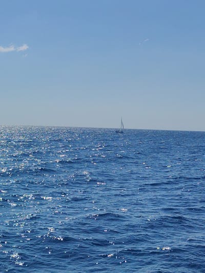 A sailboat with a white sail is visible on the horizon, set against a clear blue sky and a vast expanse of shimmering blue ocean. The sun reflects brightly off the water, creating a sparkling effect. A small, wispy cloud is present in the upper left portion of the sky. The scene appears to be a tranquil day at sea, possibly near the coast of Sipias Municipal Unit, Greece, known for its scenic maritime landscapes.