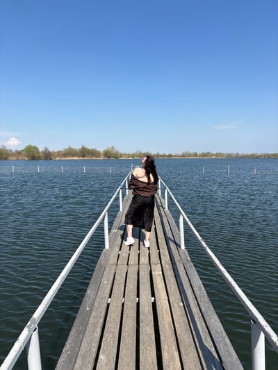 The photograph, taken in Crokhovo, Russia, presents a woman on a wooden dock extending into a body of water. The dock is constructed of parallel wooden planks and has white metal railings. The woman is facing towards the end of the dock, resting her hands on the railing. She is wearing a dark dress and a dark-colored jacket, and white sneakers. The body of water is dark blue, with small white posts protruding from its surface. A line of trees is visible on the opposite bank. The sky is clear blue. The photograph appears to be taken on a sunny day.