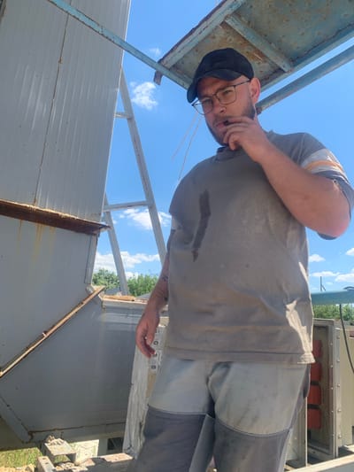 This media file shows a person in an outdoor, industrial setting. The person, who appears to be a man, is wearing a grey t-shirt with horizontal stripes on the shoulder, dark pants, and a black cap. He is wearing glasses and has his hand near his face, touching his mouth. The sky above is blue with a few white clouds, suggesting it is daytime and likely fair weather. The background features metal structures, including what looks like a large, corrugated metal wall or roof section and some rusty metal beams. There are also some trees visible in the distance. The scene appears to be one of manual labor or a work environment, possibly related to construction or maintenance. The orientation of the image is unusual, appearing as if taken from a low angle and rotated. The man's expression is difficult to ascertain clearly due to the angle and his hand, but he does not appear to be smiling. The overall impression is one of a candid, unposed moment captured while working outdoors. There is no visible text in the image.