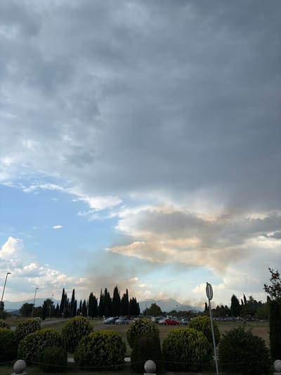 The image depicts a wide, open area on the outskirts of Golubovci, Montenegro, under a dramatic sky filled with a mix of dark storm clouds and lighter, scattered clouds. In the distance, a plume of smoke rises from behind a line of trees and mountains, suggesting a fire or other atmospheric disturbance. The foreground features a manicured area with rounded, lush green bushes and decorative spherical stone elements. Beyond this, a paved parking lot is visible, occupied by several cars of various colors. A row of tall, slender cypress trees stands between the parking lot and the distant landscape. The overall impression is one of a rural or semi-rural setting experiencing a change in weather or an environmental event.
