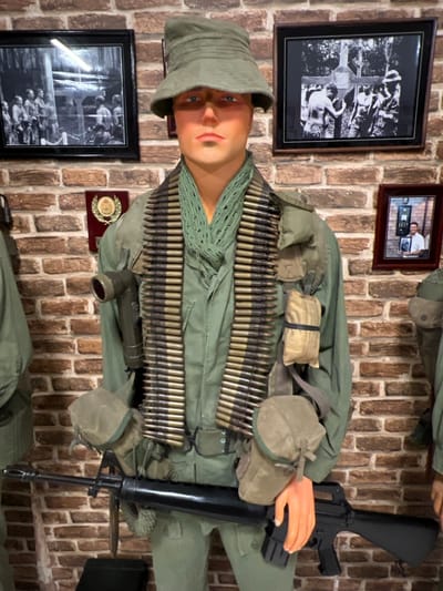 A mannequin dressed in a green military uniform stands against a brick wall. The mannequin is wearing a bucket hat and has ammunition belts draped across its chest. It holds a black rifle. Behind the mannequin are three framed pictures and a small medal.