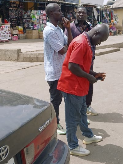 The image depicts three adult males standing on a paved surface next to a parked dark-colored Toyota Camry sedan in Kadoma, Zimbabwe, under bright daylight.

In the immediate foreground, the rear of a dark grey Toyota Camry, displaying its emblem, is visible on the left. To the right of the car stands an adult male, bald, wearing a red short-sleeved polo shirt, blue jeans, and light-colored sneakers. He is looking downwards, holding a black smartphone with both hands.

Behind him and slightly to the left stands a second adult male, bald, wearing a light blue and white checkered short-sleeved shirt, dark trousers, and light green sneakers. He has his right hand raised towards his mouth area and is looking towards the left side of the frame.

Further back and to the right of the second male is a third adult male, bald, wearing a dark patterned short-sleeved shirt, dark trousers, and light-colored sneakers. He is looking generally forward or slightly to the right.

The background features a commercial street scene with a storefront displaying various merchandise, including packaged goods and clothing, under an overhang. Boxes are stacked on the ground. A concrete curb separates the immediate foreground from the background structures. Strong shadows indicate sunny conditions.