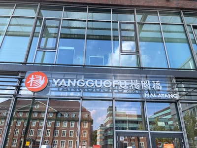 The image shows the exterior of a YANGGUOFU MALATANG restaurant in Bottrop, Germany. The restaurant has a modern facade with large glass windows and brick accents. A prominent orange circular logo with a white stylized bird symbol is visible on the left side of the building, next to the restaurant's name written in both English and Chinese characters. Below the name, the word "MALATANG" is displayed in English. The reflection in the windows shows an older brick building with many windows across the street, as well as some modern buildings and trees. A black railing runs along the bottom of the visible facade.