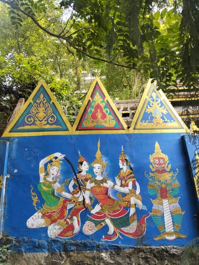 The image shows a vibrant mural on a blue wall, featuring figures in traditional Southeast Asian attire. The mural is framed by three triangular motifs at the top, each adorned with gold patterns against green and red backgrounds, depicting seated figures. Below these, a group of dancers and a large, imposing figure are painted. Four female dancers, adorned in ornate headdresses, gold jewelry, and brightly colored skirts and tops, are depicted in dynamic poses, suggesting movement and celebration. One dancer has her arms outstretched, while others are shown in more elaborate stances. To the right, a tall, fierce-looking character, possibly a demon or guardian from mythology, stands prominently. This figure is dressed in a green and gold costume with red accents and has a striking red face with wide eyes and a gaping mouth.

The mural is set outdoors, with lush green trees and foliage visible above the wall, suggesting it is daytime. Sunlight filters through the leaves, casting dappled light on the scene. The wall itself appears weathered, with some chips and cracks visible, particularly along the base, which is made of rough stone. The overall scene conveys a sense of cultural artistry and traditional storytelling, possibly from a temple or a public space in Gudangkahuripan, Indonesia, given the location context. There is no visible text in the image.