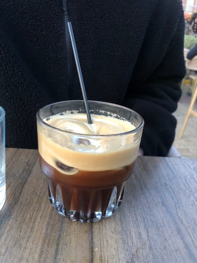 The image shows a glass of iced coffee on a wooden table. There is a black metal straw in the coffee. The coffee is made up of two layers: a darker coffee layer on the bottom and a lighter layer of milk foam on top. The glass has a fluted bottom and is sitting on a table with a wooden surface. The table is brown and has a slightly rough texture. The coffee is partially obscured by a black jacket, which is partially visible in the upper left corner of the image. The location is a cafe in Palaio Faliro, Greece, based on the given latitude and longitude.  There is no person visible in the image.  It is a still-life.  The person's arm is visible in the background.  The camera is at a slightly low angle and the subject is in focus.  The image is well-lit.