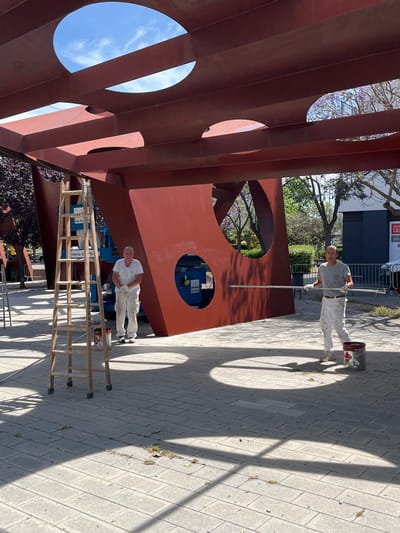 Two individuals, dressed in white work clothes, are engaged in painting a large, red, metal structure. The structure appears to be an abstract sculpture or architectural element with large circular openings. A wooden ladder stands to the left of the frame, near a bucket of paint. Another bucket of paint is positioned near the person on the right, who is holding a long roller on an extension pole. The scene is outdoors, on a paved area with sunlight casting shadows. In the background, trees with purple blossoms are visible, suggesting a park or public space. A blue hydraulic lift is also partially visible behind the sculpture. The overall impression is one of ongoing maintenance or renovation work on a public art installation. The location is identified as Sant Boi De Llobregat, Spain.
