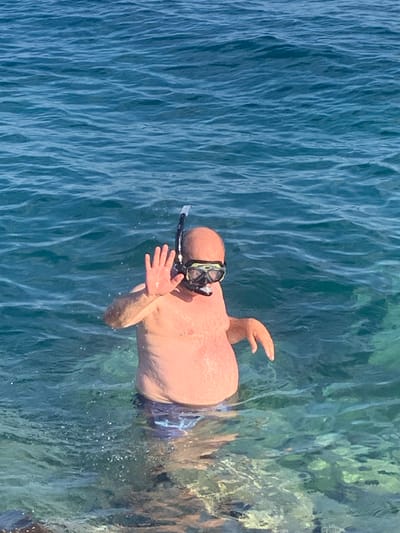 The image shows a man with a snorkel and mask in the clear blue waters of the Mediterranean Sea, likely near the coast of Orbetello, Italy. The man is shirtless and wearing swim trunks. He is facing the camera and waving with his right hand. The water is calm, with gentle waves and sunlight reflecting off the surface. The seabed is visible in some areas, with rocks and aquatic plants. The overall scene evokes a sense of relaxation and enjoyment of a summer day at the beach.