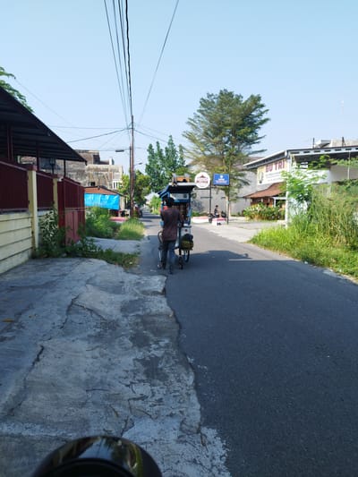 In Kricak, Indonesia, a man is walking down a paved road, pushing a cart with a covered structure. He is wearing a dark brown t-shirt and dark pants. The cart appears to be a mobile food vendor, with a blue and white canopy. To the left of the road is a sidewalk with cracked concrete and overgrown vegetation. Beyond the sidewalk, there are buildings with a red fence and a metal roofed structure. Further down the road, on the right side, is a building with a sign that reads "BU TRI". The sky is clear and blue, and there are power lines running overhead. The scene depicts a typical street in a residential or commercial area.