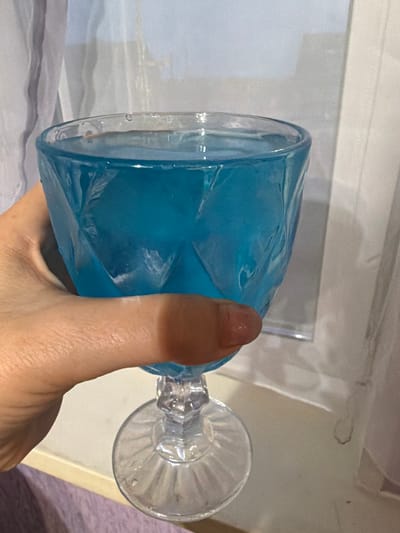 A hand is holding a stemmed glass filled with blue liquid. The glass has a faceted design. The background consists of sheer white curtains and a light-colored surface, possibly a windowsill. The overall scene is indoors, with soft lighting. There is no information about the location being the city of Нуркеево, Russia in the image itself.