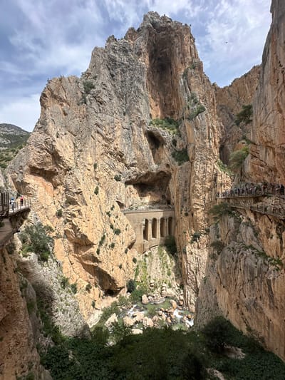 This vertical image depicts a dramatic view inside a deep gorge, characteristic of the Caminito del Rey in Ardales, Spain. Towering, rugged rock walls, composed of stratified layers in shades of light brown and gray, dominate the scene, rising steeply towards a partly cloudy sky. Sparse green vegetation clings to crevices and ledges on the rock faces.

A river with turbulent, light green water flows along the bottom of the gorge, winding around numerous rocks and boulders. Man-made structures are prominent features, showcasing impressive engineering. An elevated walkway, constructed from wooden planks and metal supports with railings, traverses high along the rock face. Numerous small figures, people walking along this path, are visible, highlighting the scale of the canyon.

Further down and to the right, old concrete or stone structures with arched openings are built directly into the rock, possibly remnants of an old aqueduct or tunnel. Another section of a wooden pathway is visible at a lower elevation in the foreground on the right, also with a few people on it.

The lighting suggests it is daytime, possibly morning or afternoon, with indirect sunlight illuminating the rock surfaces. The scene conveys a sense of awe, adventure, and the grandeur of the natural landscape combined with human ingenuity. No discernible text is visible in the image.
