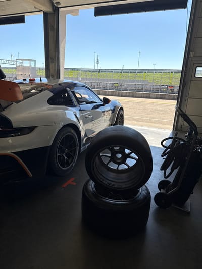 The image depicts a pit garage at the Sachsenring race circuit in Hohenstein-Ernstthal, Germany. The view is from inside the garage looking out towards the racetrack under a clear sky.
In the foreground, slightly right of center, two black racing tires with multi-spoke rims are stacked on the dark concrete floor; the upper tire is inverted. To the left, a white, black, and orange Porsche 911 GT3 race car is partially visible, parked with its rear section within the garage bay and its front end oriented towards the track. The car displays the number '25' and "SenseL" branding on its side, and features a prominent orange rear wing. A red 'T' marking is present on the garage floor near the car's rear wheel. On the far right, a dark, wheeled equipment cart is partially visible.
Beyond the open garage bay, the asphalt track surface is visible. In the background, a low concrete barrier separates the track from a tall green safety fence, behind which green foliage and several distant utility poles are discernible. No individuals are present in the scene.