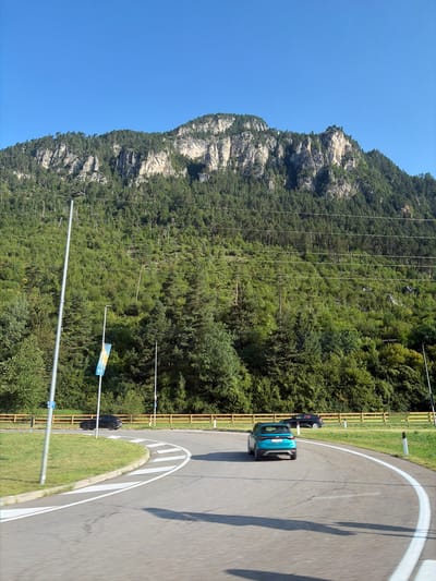 The image shows a winding road in the city of Ziano di Fiemme, Italy, with a steep, tree-covered mountain rising in the background under a clear blue sky. A teal-colored car is driving on the road, with motion blur indicating it is moving. To the left of the road, there is a grassy area with a streetlamp and a white pole with a blue and yellow flag. Another black car is parked further down the road behind a wooden fence. The mountain slope is densely covered with green trees, and rocky outcrops are visible towards the summit. Power lines stretch across the mountain. The overall scene depicts a scenic road through a mountainous, forested landscape.