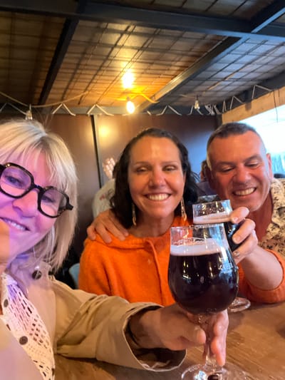 This candid, rotated photograph captures three smiling individuals enjoying a social gathering indoors, likely in a restaurant or bar in Saint-Priest-en-Jarez, France. The warm, artificial lighting suggests it's evening.
In the foreground, a woman with blonde hair and black-rimmed glasses smiles broadly at the camera. Her face is illuminated with a soft purple glow. She wears a light-colored top with a visible crocheted or patterned detail.
Behind her, a woman with dark hair, dressed in a vibrant orange sweater, also smiles. Her left hand rests on the shoulder of the blonde woman, while her right hand holds a stemmed glass filled with a dark liquid, possibly red wine or dark beer. She appears to be interacting with the bottle.
Further back, partially obscured by the middle woman, a man with short dark hair, wearing a patterned shirt over an orange undershirt, grins widely. His hands are positioned near a dark bottle, from which the drink is being poured into the glass held by the woman in orange.
The immediate setting includes a wooden table where the drinks are placed. The background features distinct wooden wall panels arranged in a grid pattern, illuminated by bright, exposed light fixtures, suggesting an industrial or rustic-modern interior design. Some metallic structures and wires are also visible.
All three subjects appear happy and relaxed, indicated by their wide smiles, suggesting an enjoyable social event. No clear, readable text is visible on the bottle label or any signs in the background.