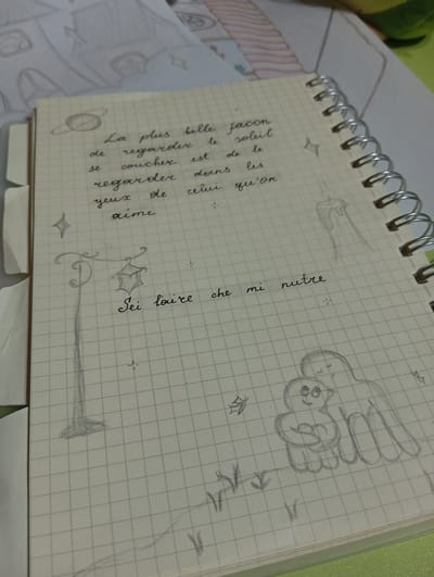 The image is a close-up of a notebook with graph paper. The notebook contains handwritten text in French and pencil sketches. The French text reads: "La plus belle façon de regarder le soleil se coucher est de le regarder dans les yeux de celui qu'on aime." This translates to "The most beautiful way to watch the sunset is to watch it in the eyes of the one you love."
The sketches include a planet with rings, a street lamp, a faint drawing of a candle, two figures embracing, and some small plants or grass. The notebook is open and appears to be a personal journal or sketchbook. The background shows another piece of paper with faint sketches and a green surface. There is no indication of the location being Kokand City, Uzbekistan.