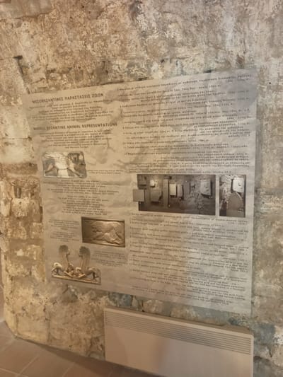 The image shows a section of a wall with a large informational poster in the center. The poster is mounted on the rough, stone wall of an ancient structure. The poster is divided into sections with Greek and English text and includes several images of ancient art. The text describes "MIDDLE BYZANTINE ANIMAL REPRESENTATIONS" and mentions their discovery in the region of Chalkis. The poster's content suggests the location is within the Municipal Unit of Anthidon, Greece, as it refers to discoveries in the region of Chalkis. The images on the poster depict various artistic representations of animals and mythical creatures, such as griffins, eagles, lions, and sphinxes, rendered in stone and marble, primarily in bas-relief. These are described as architectural elements or decorative items from the Middle Byzantine period. The poster also includes a quote in English and Greek. The environment appears to be an indoor space with low lighting, likely a museum or historical site, given the display of artifacts and informational material. The wall itself is constructed of large, irregular stones, indicative of ancient architecture.