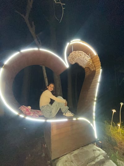 A young woman hangs upside down from a light-lined, wooden structure at night. The structure, which resembles an ampersand and is adorned with carved wooden details at the top, is located in Sungurlare, Bulgaria. The woman is wearing a light-colored shirt, light-wash jeans, and white shoes, and she appears to be holding onto the structure. A pink and white patterned fabric hangs down behind her. In the background, there are illuminated mushroom-shaped lights, trees, and a stone structure. The area is dark, illuminated primarily by the structure's lights.