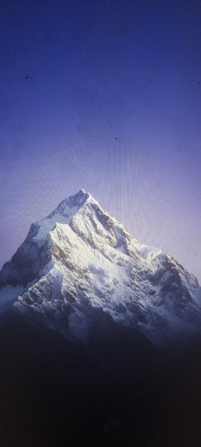 A majestic, snow-capped mountain dominates the frame, its peak reaching towards a clear, deep blue sky. The mountain's slopes are rugged and textured, with patches of snow highlighting its sharp ridges and deep crevasses. Sunlight bathes the upper portion of the mountain, creating a dramatic contrast between the illuminated snow and the shadowed valleys below. The lower part of the mountain fades into darkness, obscuring the base. The image appears to be a photograph or digital rendering. There is no indication of a city location, including Kudymkar, Russia, within the visual content.