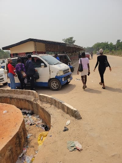 This image captures an outdoor scene in the city of Candelaria, Colombia, under a hazy, light-gray sky. A white and blue mini-van, bearing the text "TRANSCARGA" on its side, is positioned on a dirt road. Four individuals are gathered near the passenger side of the van; one woman in a red top and blue jeans stands closest to the vehicle, another woman in a purple patterned top is partially obscured near the front passenger door, a person in dark clothing is partially within the open front passenger door, and a male in a blue shirt is near the rear passenger door.

To the right of the van, two women walk away from the scene along the dirt road. The lead woman wears a light pink t-shirt and white skirt, carries a dark shoulder bag, and holds a phone in her right hand. She is followed by another woman dressed in a black long-sleeved top and black pants, with a patterned headscarf. Further down the dirt road, several indistinct figures and vehicles, including a red car and a white car, are visible.

In the mid-ground, a long, open-sided shelter with a dark roof and a light-colored base runs parallel to the road. An auto-rickshaw is partially visible beneath the shelter. The foreground features a curved concrete drainage ditch, which is heavily filled with various types of refuse, including plastic bottles, bags, and fabric scraps. Additional litter is scattered on the surrounding dirt ground. Sparse green vegetation is present in the distant background.