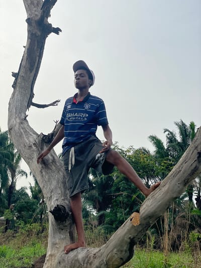 A person is lying horizontally on a large, fallen tree in Agba Umana, Nigeria. The person is wearing a blue and white striped shirt, gray shorts, and a baseball cap. Their bare right foot is resting on the tree trunk. The tree is positioned in a natural environment with other trees and vegetation visible in the background. The sky is overcast.