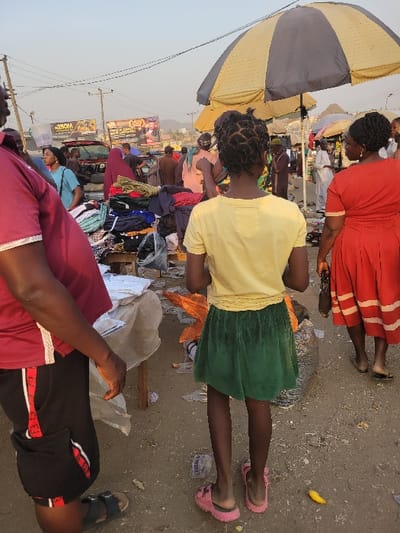 A busy outdoor market scene. People browse through piles of clothes spread on tables and the ground. One individual in a red dress and another in a yellow top and green skirt stand out. An umbrella offers shade, and various advertisements adorn the background. The ground appears to be a mix of dirt and scattered debris. Footwear ranges from sandals to closed shoes, adding to the lively atmosphere of the market.